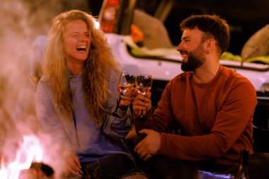 Two friends sit by a warm campfire under the stars, sharing joyful laughter while raising their glasses. The atmosphere is relaxed, with glowing flames and a serene backdrop.
