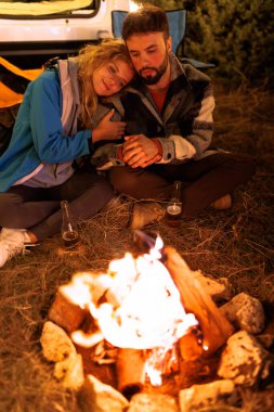 A couple sits close together by a warm campfire, sharing a peaceful moment. They appear relaxed while enjoying the beauty of nature at night. Their surroundings are calm and serene.