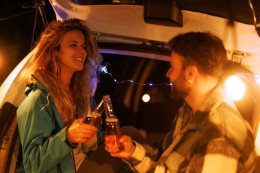 Two people share a joyful moment, raising their drinks while seated in a car trunk. The night sky and decorative lights create a warm, relaxed atmosphere perfect for socializing.