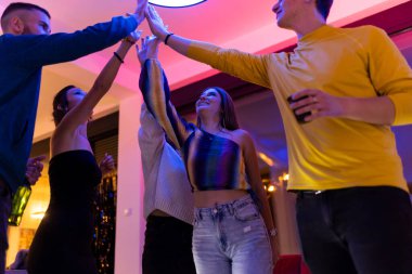 A cheerful group of friends gathers at an indoor party, engaging joyfully, and sharing the moment by giving high fives. The festive environment is enhanced by colorful lighting,