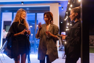 Three women are sharing a casual conversation while holding drink glasses at a stylish outdoor setting adorned with string lights during the evening.
