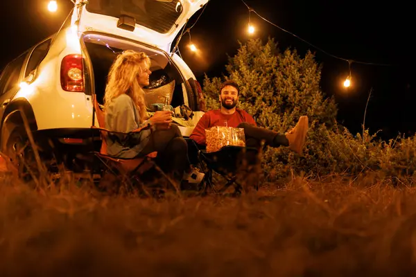Two friends relax in camping chairs near their car at night. They share snacks and laughter under string lights, enjoying the peaceful outdoor atmosphere.