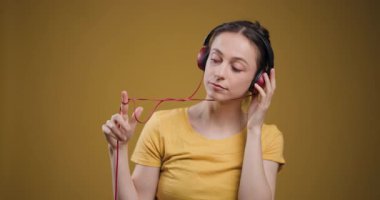 A serene young adult woman closes her eyes while listening to music through red headphones against a plain yellow backdrop.