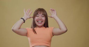 A joyful Asian woman laughing with her arms raised in a moment of happiness, captured in vibrant clothing against a plain background.