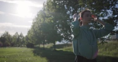 A young woman in a light green jacket stretches her arms behind her head in a peaceful park setting during sunset, portraying relaxation and fitness.