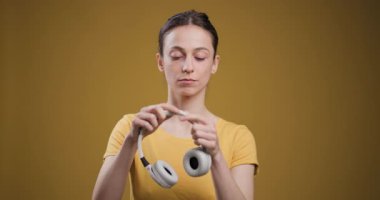 A serene young woman with closed eyes, wearing a yellow t-shirt, listens to music on white wireless headphones against a yellow backdrop.
