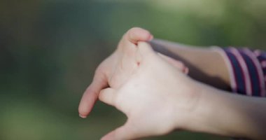 A woman is engaging in a stretching routine, focusing on her hands in a calm outdoor location surrounded by greenery. The atmosphere is peaceful and refreshing.
