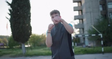 A young man is preparing for a basketball practice on an outdoor court in the city during sunset. He adjusts his gear with enthusiasm, ready to engage in some fun.