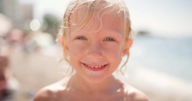 Close-up of a delightful little blonde girl with wet, curly hair laughing and making funny faces on a sun-drenched beach during a joyful summer vacation, capturing pure childhood happiness.