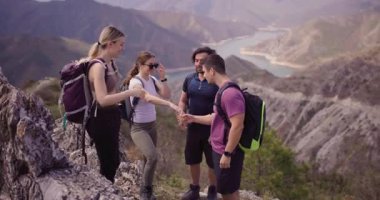 Dört arkadaş grubu dağlarda yürüyüş yaparken eğlenceli bir anı paylaşıyorlar. Arka planda güzel bir göl ve dağlarla beşlik çakıyorlar..