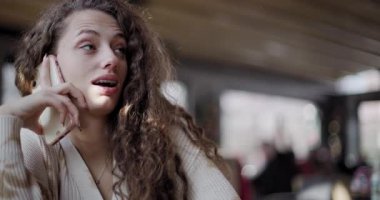 A young curly-haired woman with braces looks puzzled and concerned as she talks on her phone in a public space.