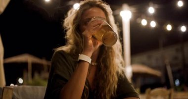 A woman sits at a restaurant table in the evening, savoring a cold beer under twinkling lights, enjoying the cozy atmosphere of a night out.