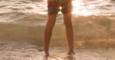 Golden light illuminates a persons bare legs as gentle ocean waves wash over their feet on a sandy beach during a beautiful sunset or sunrise.