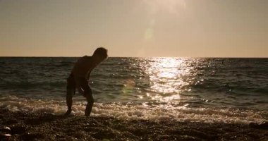 A young boys silhouette is captured as he playfully throws rocks into the calm ocean waves at sunset, with golden sunlight reflecting beautifully on the water during a peaceful summer evening on the