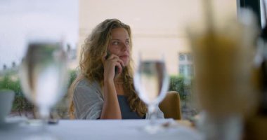 A woman enjoys a meal at a restaurant, sitting by the window while engaged in a phone conversation. The ambiance features scenic views and a rainy backdrop, adding a cozy atmosphere.