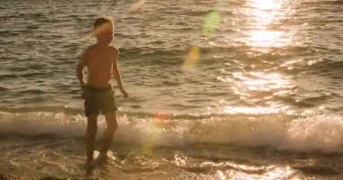 A young boy joyfully plays in the shallow waves of a beautiful beach during a golden sunset, with the suns reflection sparkling brightly on the waters surface.