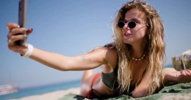 A person is lying on a beach blanket, taking a selfie while enjoying the sun and clear sky. The beach is lively with a relaxing atmosphere and gentle waves in the background.