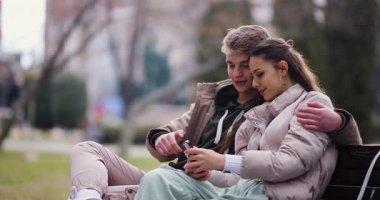 Affectionate young couple enjoying a cozy moment on a bench, taking a selfie with a smartphone, surrounded by a calm urban park setting.