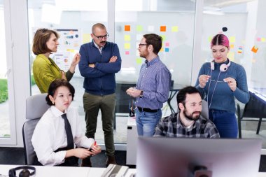 A group of diverse businesspeople discuss strategies and share ideas in a modern office. Some are seated while others engage in conversation and collaboration, creating a dynamic atmosphere.