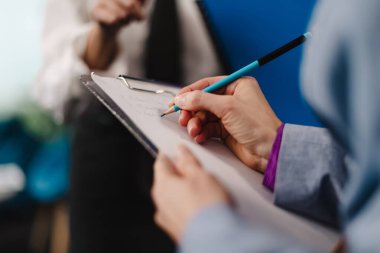 Two business professionals are engaged in a discussion in a modern office. One individual is taking notes on a clipboard while another gestures, sharing ideas.