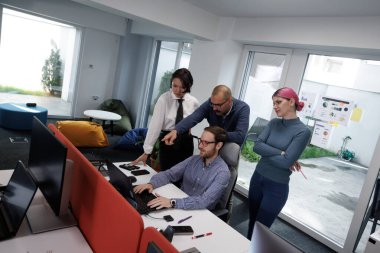 A group of four business professionals works together at a workstation in a bright and contemporary office. They are engaged in a discussion about a project, focusing on a computer screen.