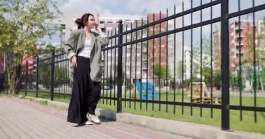 A young Kazakh woman engages in a leisurely walk, dressed in business casual and using a smartphone, amidst an urban park setting.