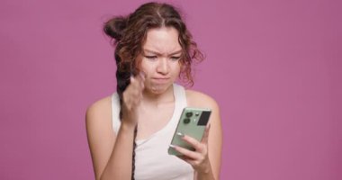 A young woman with a braided hairstyle frowns as she examines her smartphone, expressing confusion, shot against a vivid pink backdrop.