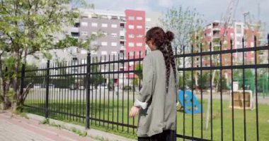 A young woman with a long braid walks by a fence in an urban park setting, clad in casual attire, depicting a relaxed outdoor activity.
