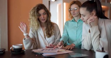 Three businesswomen engage in a productive discussion around project charts in a stylish office, sharing ideas and strategies for success while enjoying coffee.