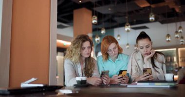 Three businesswomen gather in a modern cafe to brainstorm and exchange ideas. Engaged in discussion, they utilize their smartphones to enhance their entrepreneurial efforts over coffee.