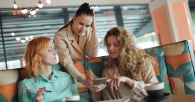 Three businesswomen gather in a trendy cafe, sharing ideas and insights while reviewing information on a mobile device. The atmosphere is vibrant and collaborative.