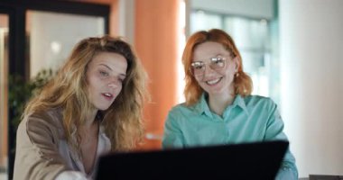 Two professional women engage in a productive discussion while examining important documents on a laptop in a modern office. Their focus reveals a commitment to achieving their business goals.
