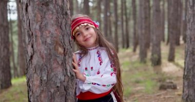 Happy smiling Bulgarian girl in folklore costume hiding behind forest tree, Bulgaria nature