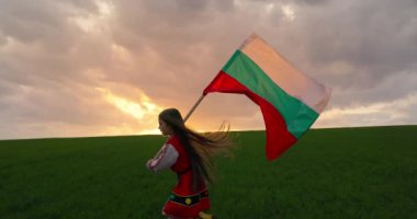 Folklor etnik kostümü içinde güzel bir Bulgar kızı ve yeşil tarım sahasında açık havada dolaşan bir Bulgar bayrağı. Özgürlük, güzellik ve Bulgar bağımsızlığı ve gurur konsepti, 4k video