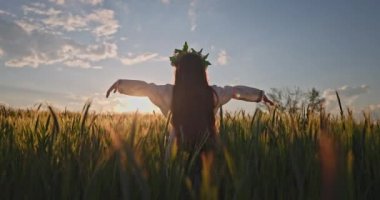 Çiçek şapkalı güzel bir genç kız, geleneksel Bulgar işlemeli etnik folklor elbisesi buğday tarlasında 4k video klibinde gün batımında