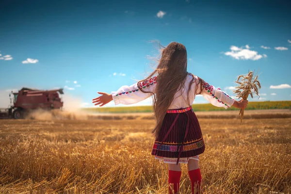 収穫時の農業の小麦畑で伝統的なブルガリアの民俗衣装を持つ若い女の子は 機械を組み合わせる — ストック写真