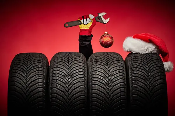 Winter car tires, big wheels, tyres, mechanic hand with wrench, christmas tree ball and Santa Claus hat on red background.