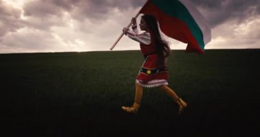 Etnik folklor kostümlü Bulgar güzel kız yeşil alanda koşuyor ve dramatik gün batımı, özgürlük, bağımsızlık ve özgürleşme Bulgaristan 'da Bulgar bayrağını gururla sallıyor