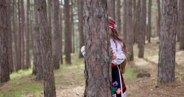 Bulgaristan 'ın dağlarındaki bir ormanda ağaç gövdesinin arkasına saklanan, etnik folklor işlemeli Bulgar kadın genç kız.