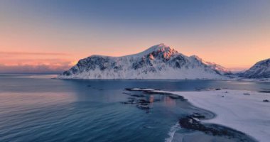 Karla kaplı dağ zirveleri, kayalık kıyı şeridi ve Uttakleiv Sahili, Lofoten Adaları, Norveç 4K video klibi üzerindeki manzaralı günbatımı.