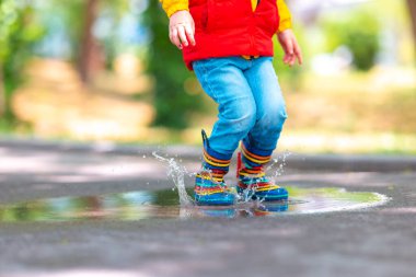 Parktaki yağmurlu su birikintisinin üzerinden atlayan renkli lastik çizmeli çocuk ayakları