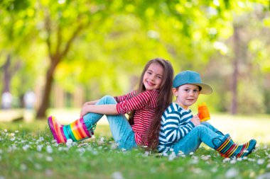 Mutlu çocuklar. Yağmurlu lastik çizmeli kız ve oğlan. Yeşil parkta, çiçek açan papatya çiçekleriyle oynuyorlar.