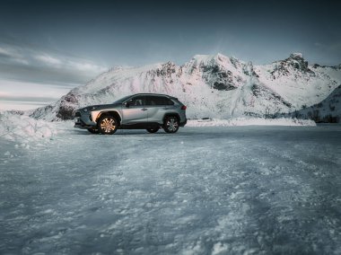 Fast modern car drives on winter road of snow and ice to snowy covered mountain peaks, scenic arctic landscape clipart