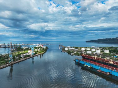 Sakin bir sahil şeridinde bulunan hareketli bir sanayi limanının panoramik görüntüsü, dramatik bulutlu gökyüzüne karşı kurulmuş gemiler, depolar ve temel tesisler sergileniyor..