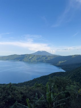 Sakin göl, parlak güneş ışığı altında yeşil tepeler tarafından kucaklanırken gökyüzünü yansıtır.