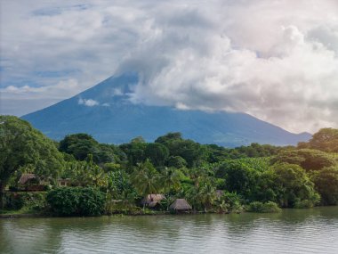 Sükûnet gölü yeşil yaprakları ve sazdan kulübeleri yansıtır. Gökyüzünde yükselen volkan ve bulutlar manzaralı bir manzara yaratır..