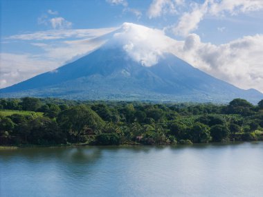 Sersemletici volkan yeşillik ve sakin suların üzerinde görkemli bir şekilde yükseliyor, berrak gökyüzünün altında doğanın güzelliğini gösteriyor..