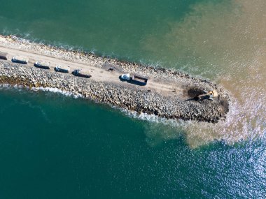Sahil iskelesinde çalışan ağır makineler ve kamyonlar gün ışığında temiz mavi sularla çevrili..