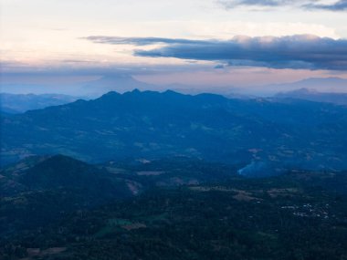 Alacakaranlık dingin dağ manzarasına yerleşir, renkli gökyüzünün altındaki uzak tepeleri ortaya çıkararak, sakin bir atmosfer yaratır..