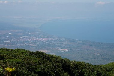 Geniş kıyı manzarası gün boyunca yemyeşil, okyanus ve uzak bir şehrin berrak bir gökyüzü altında olduğunu gösteriyor..